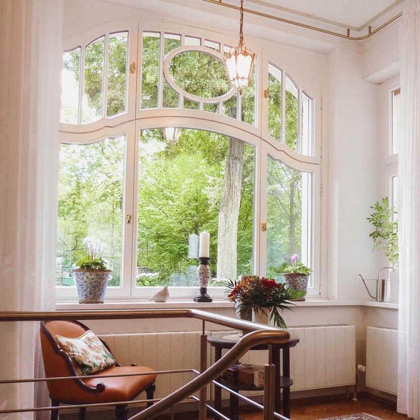 Großes Fenster mit geschwungenem Rahmen und rundem Oberlicht, davor ein Tisch mit  Sessel und Kissen