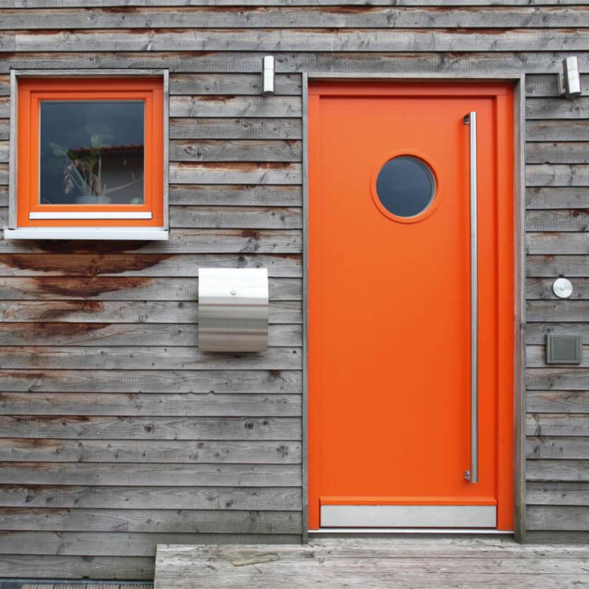 Orangefarbene Haustür mit rundem Fenster und langem Griff neben einem kleinem Fenster