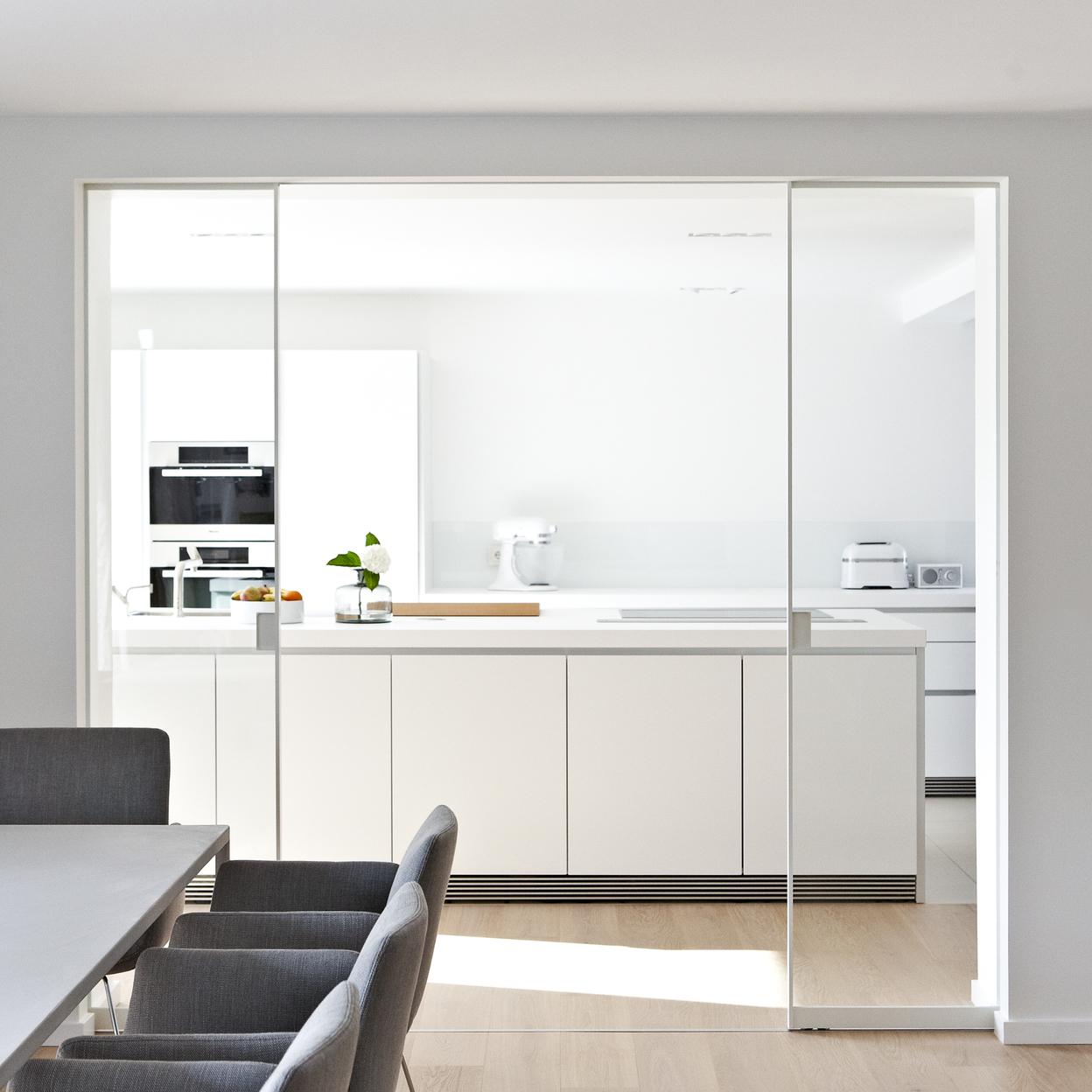 Moderne Küche und Esszimmer Eine moderne Küche und ein Esszimmer mit maßgefertigten Küchenschränken in weiß. Der Raum ist hell und minimalistisch gestaltet, mit einer großen Glas-Schiebetür zwischen Küche und Essbereich.