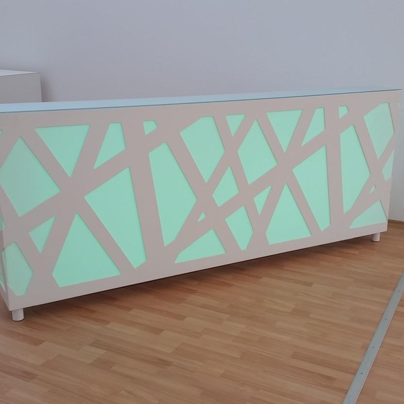 Empfangstresen mit grüner Beleuchtung Weißer Empfangstresen mit geometrischem Muster und hinterleuchteter grüner Oberfläche, aufgestellt in einem Raum mit hellem Laminatboden.