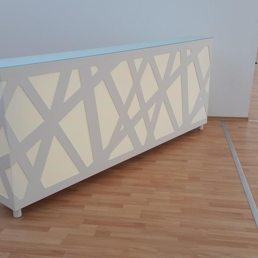 Empfangstresen mit warmer Beleuchtung Weißer Empfangstresen mit geometrischem Muster und hinterleuchteter, warmweißer Beleuchtung, aufgestellt in einem Raum mit hellem Laminatboden.