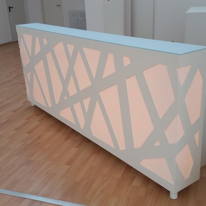Empfangstresen mit dezentem rosa Licht Weißer Empfangstresen mit geometrischem Muster und hinterleuchteter, zartrosa Beleuchtung, aufgestellt in einem Raum mit hellem Laminatboden.