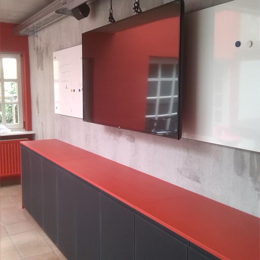 Moderne Objekteinrichtung in einem Büro, mit langen, dunklen Schränken und roter Arbeitsfläche, kombiniert mit einer roten Wanddekoration. Effektiv für den professionellen Einsatz.