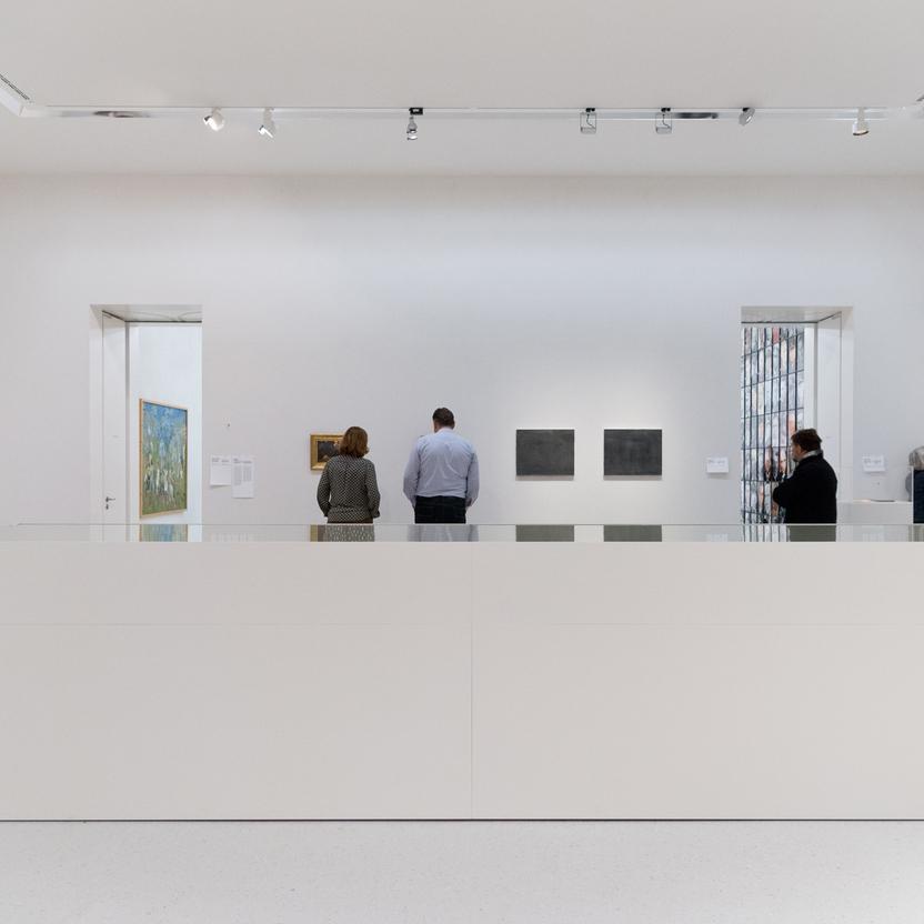 Innenansicht einer Kunstgalerie mit Besuchern, die Gemälde betrachten. Zentral steht ein großer, weißer Vitrinenbau, der Kunstwerke präsentiert.