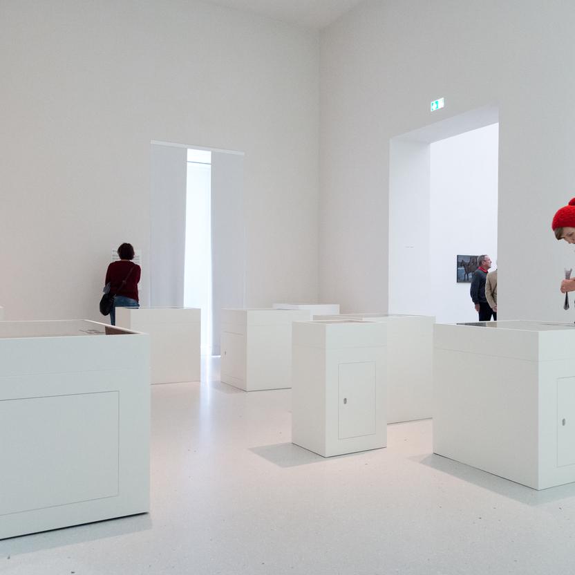 Moderne Kunstgalerie mit Besuchern und mehreren minimalistischen Vitrinenbauten, die in einem hellen, offenen Raum verteilt sind.