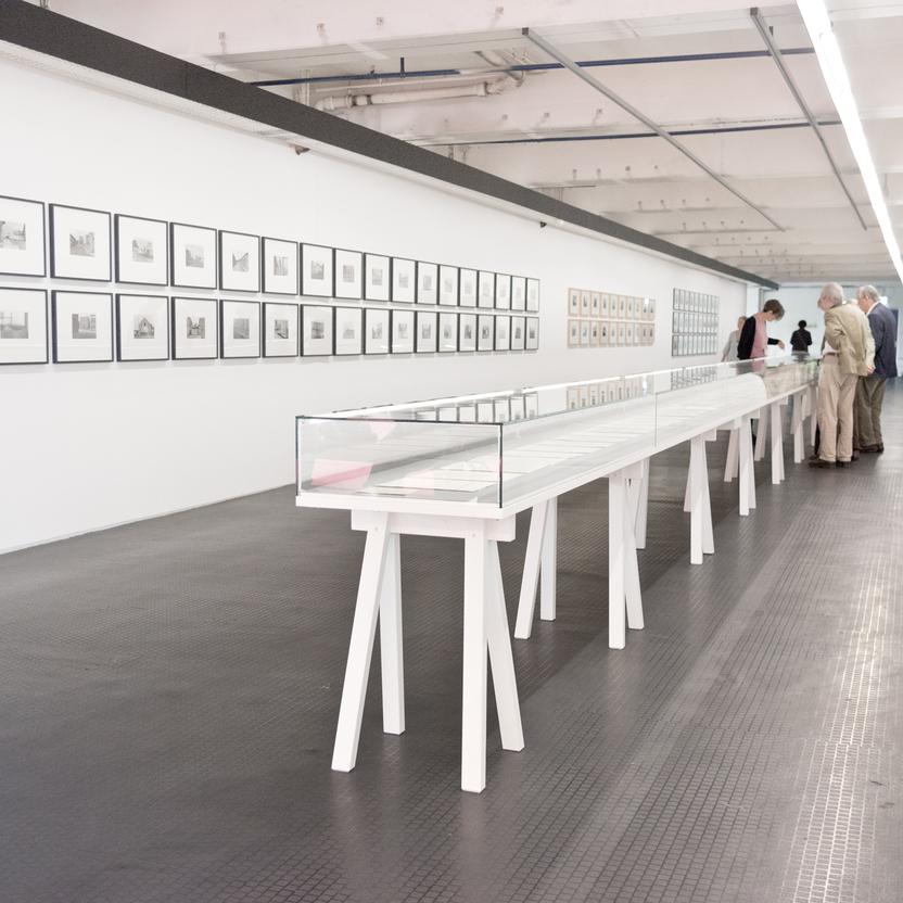 Eine lange Glasvitrine auf weißen Gestellen steht in einem hellen Ausstellungsraum. An der Wand hängen zahlreiche gerahmte Bilder. Besucher betrachten die Exponate.
