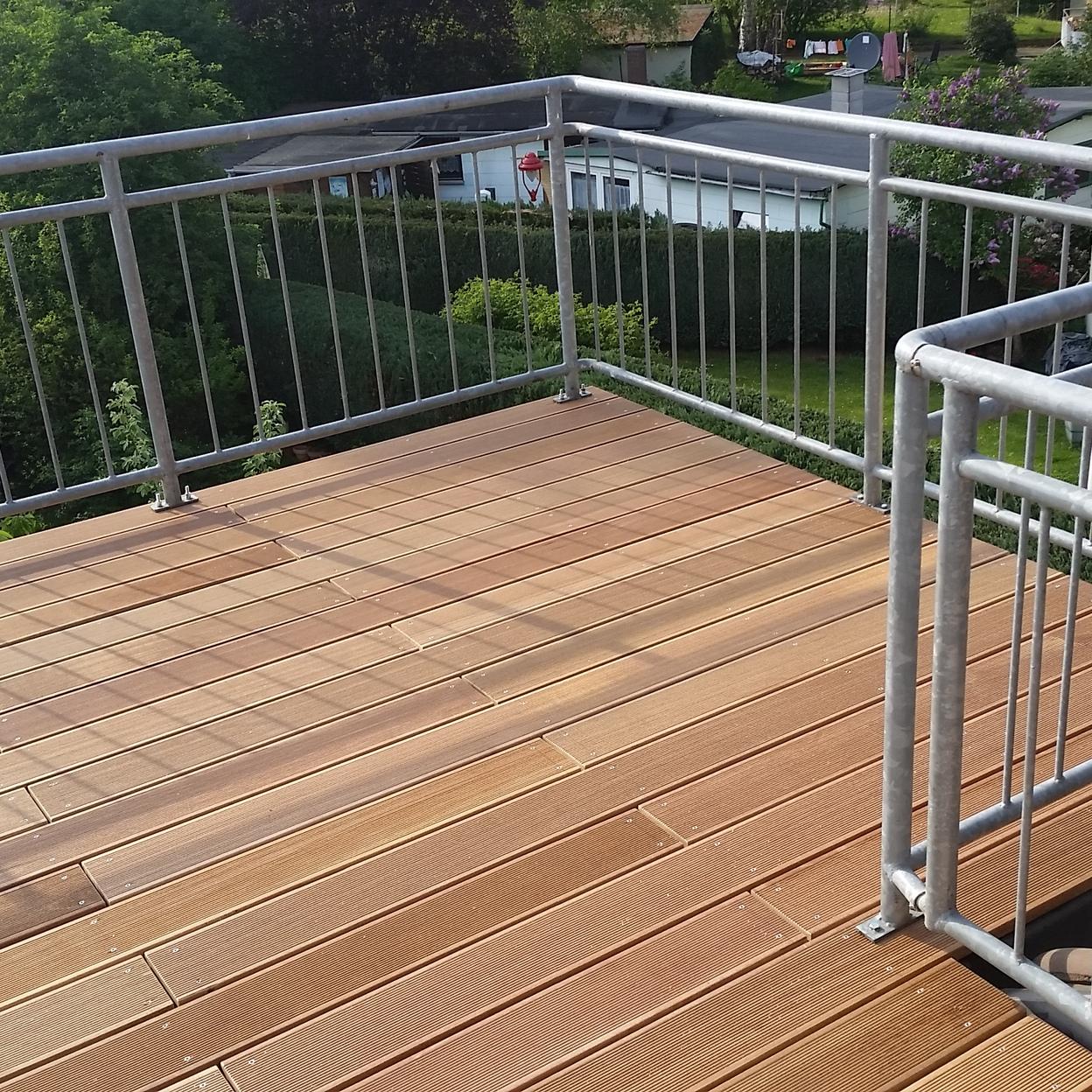 Holzterrasse mit Blick auf den Garten Eine Holzterrasse mit hellbraunen Dielen und einem Metallgeländer, umgeben von grünen Bäumen und Hecken, mit Blick auf Nachbarhäuser und Gärten. Keyword: Holzterrasse.