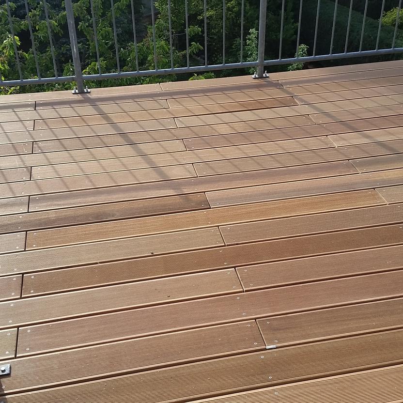 Detailansicht einer Holzterrasse mit Geländer Nahaufnahme einer Holzterrasse mit hellbraunen Dielen und einem Metallgeländer, das von Sonnenlicht beschienen wird. Im Hintergrund sind grüne Büsche sichtbar. Keyword: Holzterrasse.