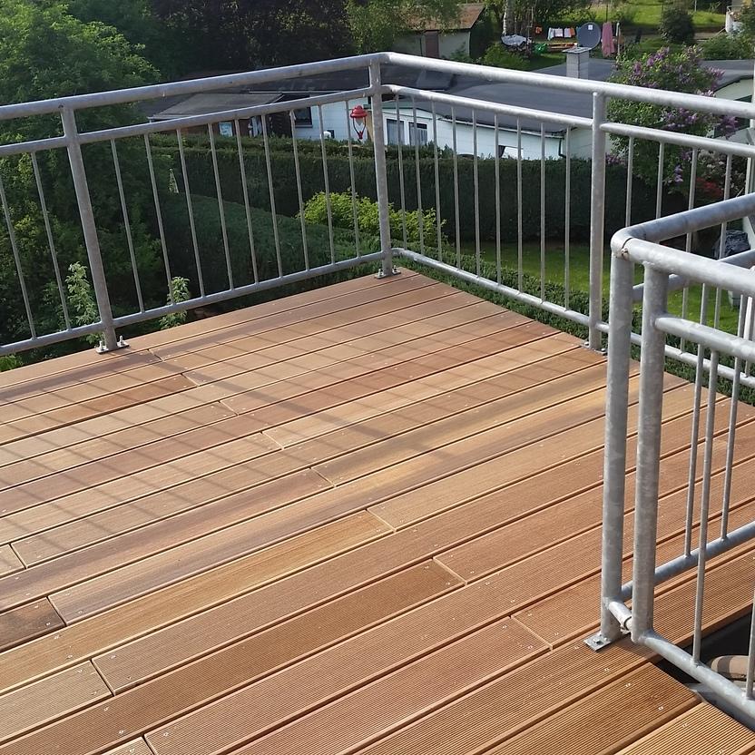 Holzterrasse mit Blick auf den Garten Eine Holzterrasse mit hellbraunen Dielen und einem Metallgeländer, umgeben von grünen Bäumen und Hecken, mit Blick auf Nachbarhäuser und Gärten. Keyword: Holzterrasse.
