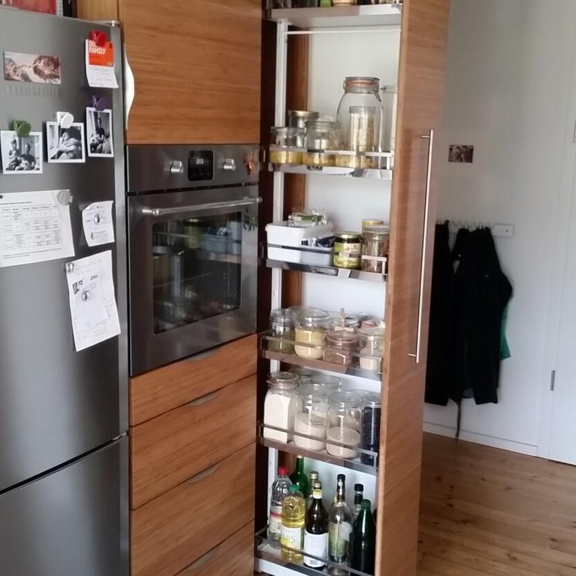 Maßgefertigte Küche mit Holzfronten, integriertem Apothekerschrank und Einbaubackofen, kombiniert mit einem modernen Kühlschrank. Der Apothekerschrank zeigt Gläser und Flaschen. Keyword: Küche nach Maß.