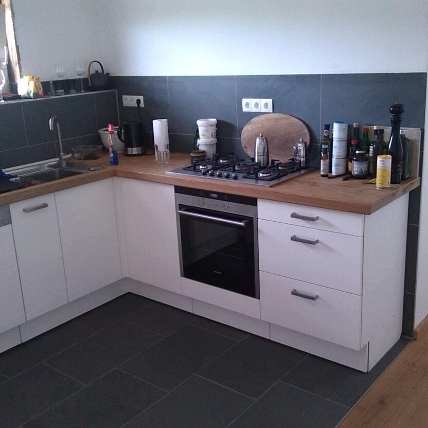 Eine L-förmige Küche mit weißen Fronten, einer Holzarbeitsplatte, eingebautem Gasherd und Spüle. Dunkle Fliesen an der Wand und am Boden ergänzen das Design.