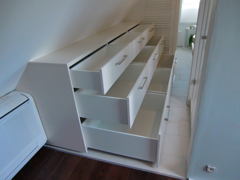 Weiße maßgefertigte Möbel mit offenen Schubladen unter einer Dachschräge neben einer Klimaanlage, auf dunklem Holzboden.