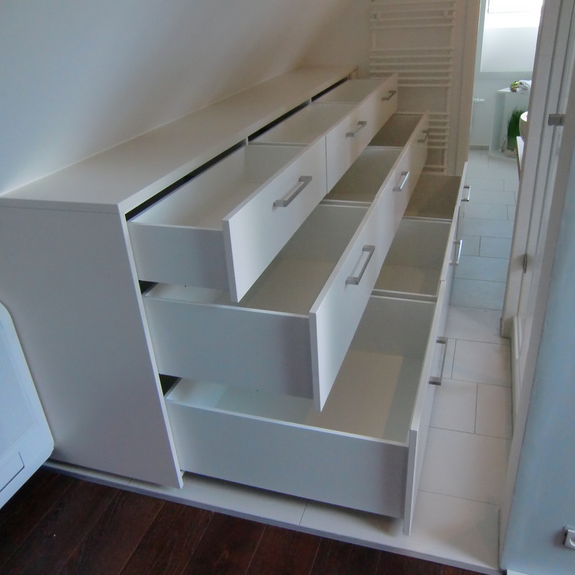 Weiße maßgefertigte Möbel mit offenen Schubladen unter einer Dachschräge neben einer Klimaanlage, auf dunklem Holzboden.