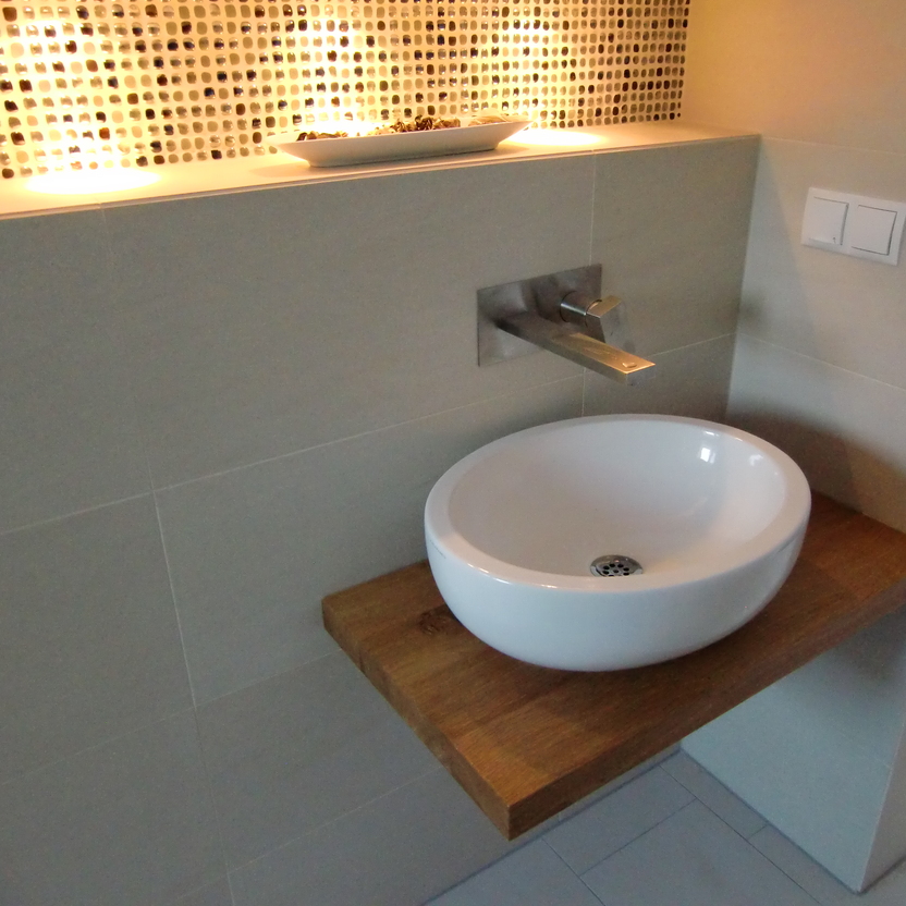 Moderne Waschschale auf einer maßgefertigten Holzplatte, montiert an einer gefliesten Wand mit einem dekorativen Mosaik als Hintergrund.