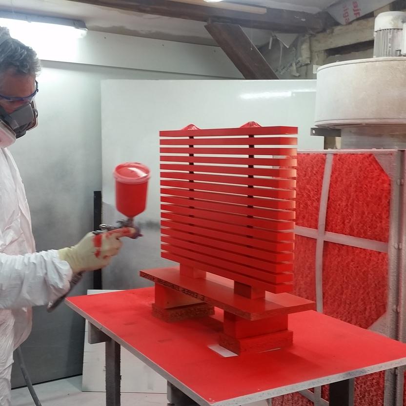 Ein Mann in Schutzkleidung lackiert Holzmöbel in leuchtendem Rot in einer professionell ausgestatteten Lackierkabine.