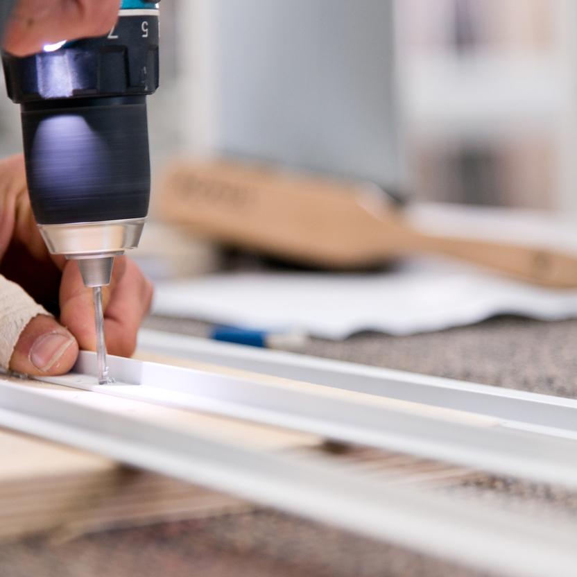 Nahaufnahme einer Hand, die mit einem Akkubohrer ein Loch in ein weißes Metallprofil bohrt. Im Hintergrund unscharfe Werkzeuge und Arbeitsmaterialien.
