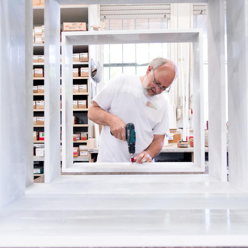 Klaus Waha arbeitet mit einem Akkubohrer an einem Rahmen in einer hellen Werkstatt, umgeben von Regalen mit Materialien und Werkzeugen.