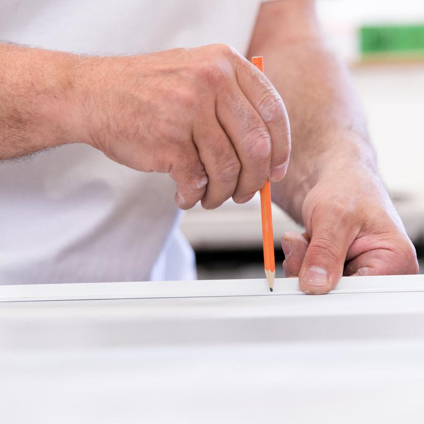 Nahaufnahme von Händen, die mit einem orangefarbenen Bleistift Markierungen auf einer weißen Oberfläche anzeichnen, um genaue Maße festzulegen.