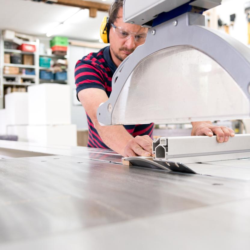 Ein Handwerker mit Schutzbrille und Gehörschutz arbeitet an einer Kreissäge, um ein Aluminiumprofil zuzuschneiden. Im Hintergrund eine gut ausgestattete Werkstatt.