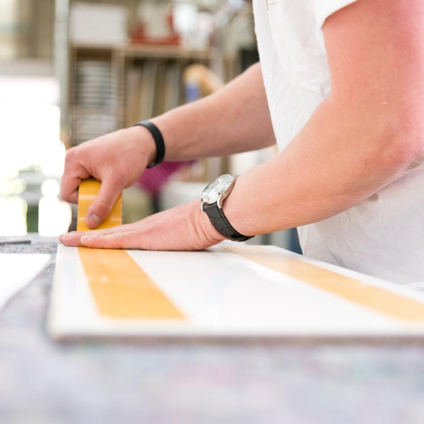 Nahaufnahme eines Handwerkers, der mit einem gelben Klebeband auf einer weißen Oberfläche arbeitet, um eine präzise Abdeckung zu erstellen. Im Hintergrund eine Werkstattumgebung.