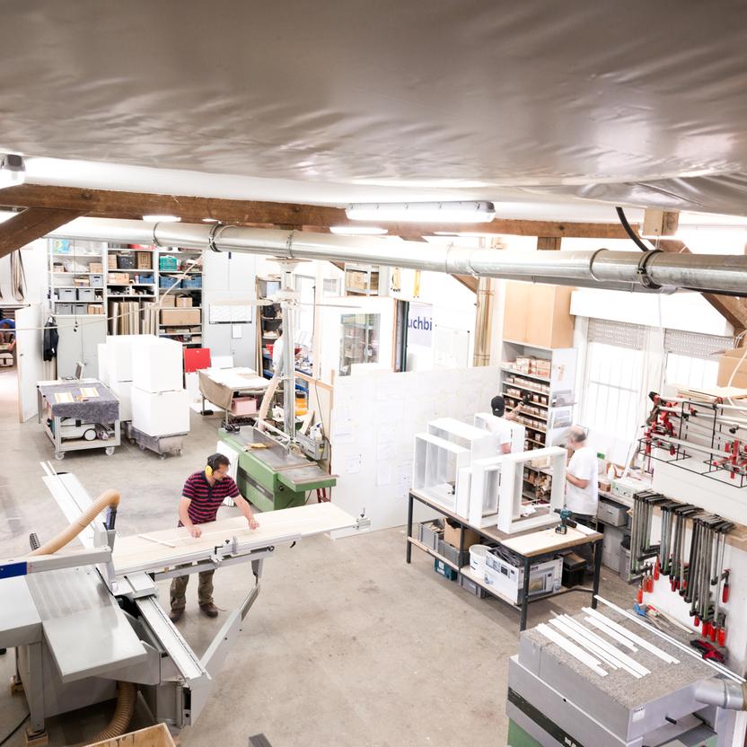 Blick von oben auf eine Schreinerwerkstatt mit mehreren Handwerkern, die an Maschinen und Werkbänken arbeiten. Die Werkstatt ist gut ausgestattet mit Werkzeugen, Maschinen und Materialien.