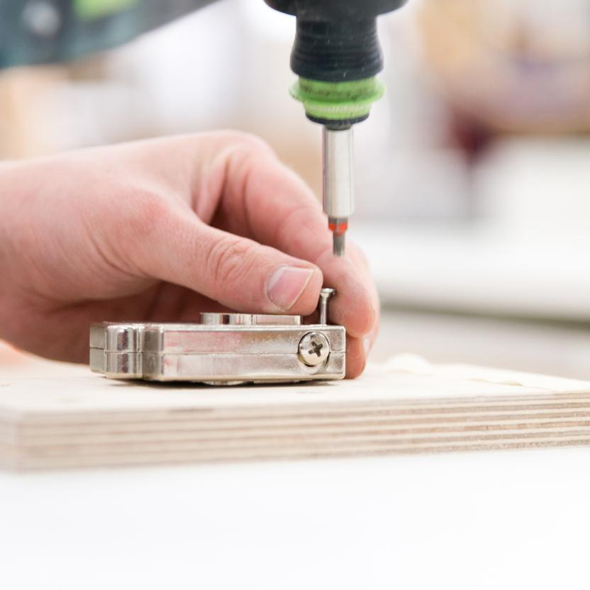 Nahaufnahme einer Hand, die ein Metallscharnier auf einer Holzplatte hält, während ein Akkuschrauber eine Schraube befestigt. Präzise Handwerksarbeit in einer Werkstatt.