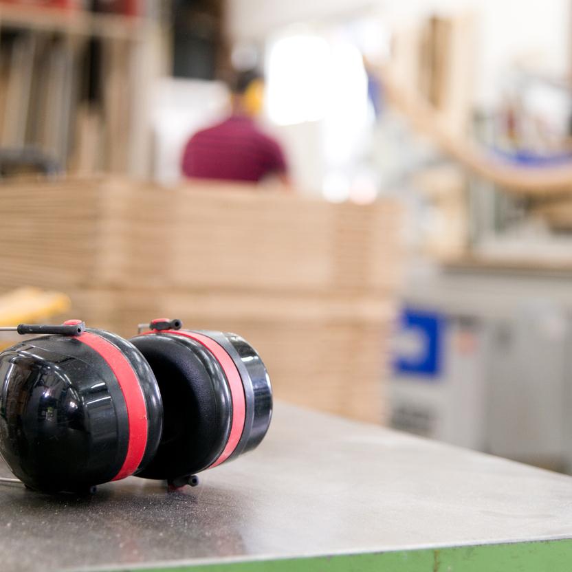 Ein schwarzer Gehörschutz mit roten Akzenten liegt auf einer Werkbank in einer Schreinerei. Im Hintergrund verschwommene Arbeitsumgebung mit Holzplatten und Maschinen.