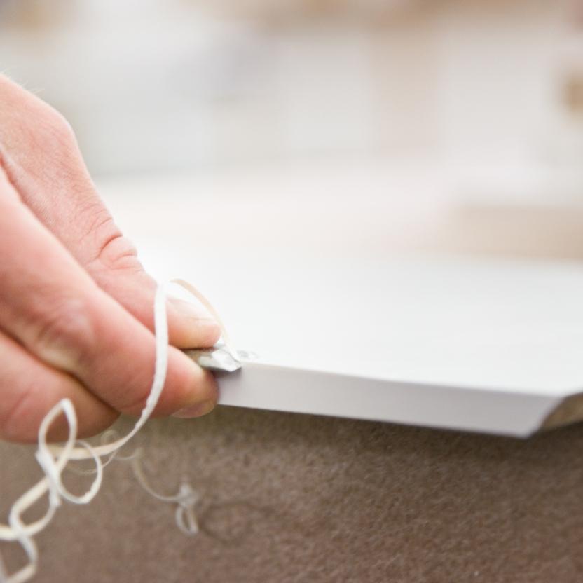 Nahaufnahme einer Hand, die mit einem Werkzeug die Kante einer weißen Platte bearbeitet. Dabei entstehen feine Späne für eine präzise Fertigung.