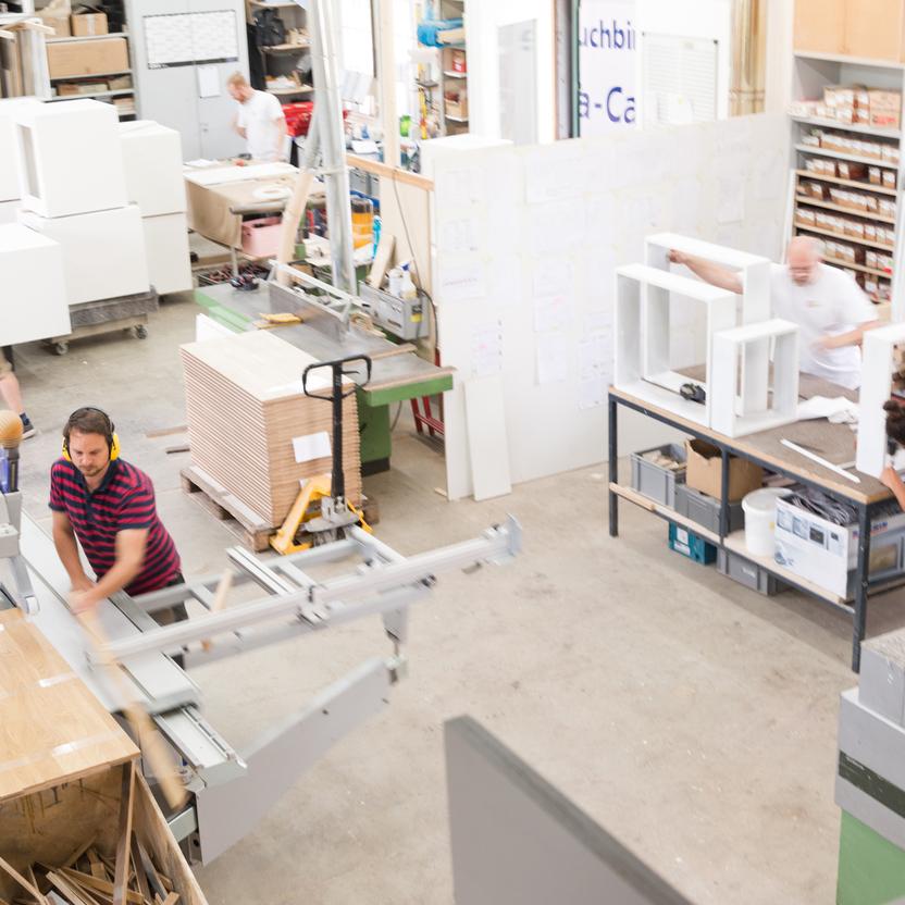 Übersicht einer Schreinerwerkstatt mit mehreren Handwerkern, die an verschiedenen Arbeitsstationen und Maschinen Holz und Möbel bearbeiten. Stapel von Materialien und fertige Module sind sichtbar.