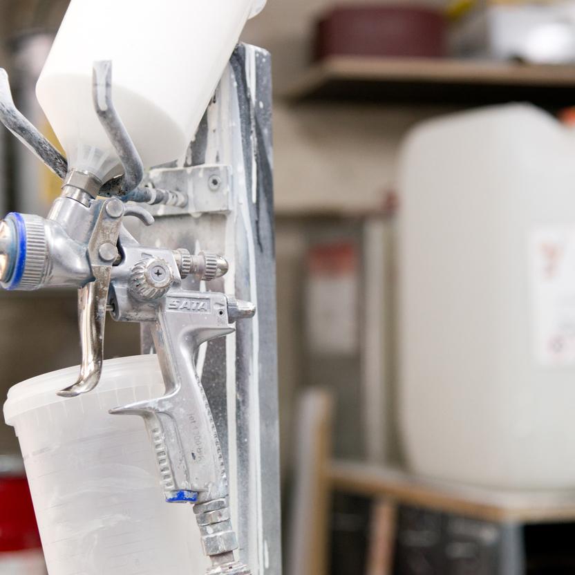 Nahaufnahme einer silbernen Spritzpistole mit weißem Behälter, montiert auf einer Halterung. Im Hintergrund stehen große Behälter und Lackiermaterialien in einer Werkstatt.