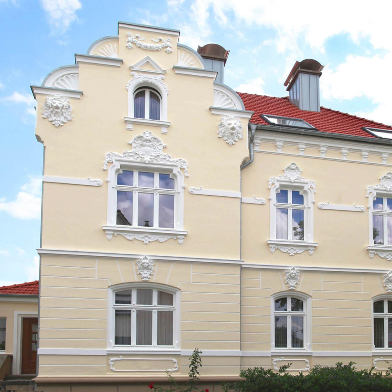 Fassade eines Hauses mit weißen Stuckverzierungen, roten Ziegeldach und mehreren Fenstern unter blauem Himmel