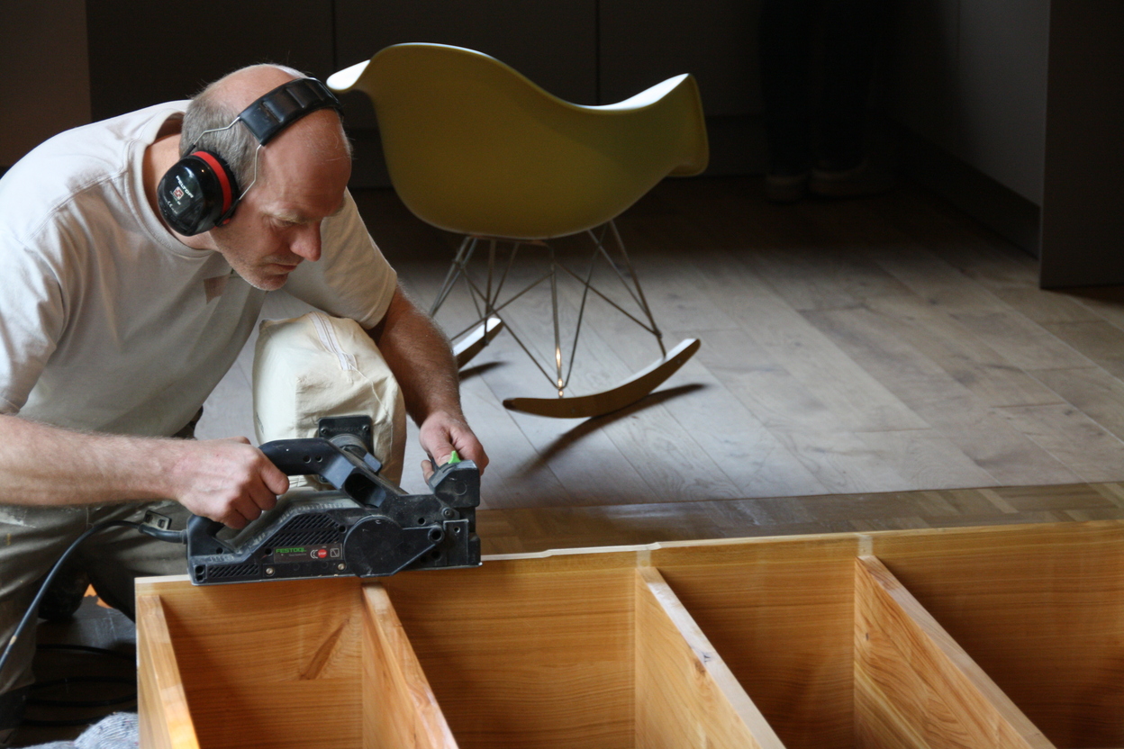 Ein Tischler bearbeitet mit einem Elektrowerkzeug ein Regal nach Maß aus Holz, während er Gehörschutz trägt. Im Hintergrund ist ein Holzboden und ein Schaukelstuhl zu sehen.