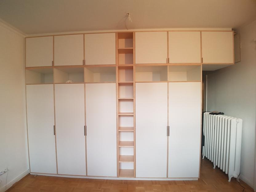 Maßgefertigter Schrank in einem hellen Raum. Ein großer, maßgefertigter, cremefarbener Schrank mit Türen und offenen Regalen in einem Zimmer mit Parkettboden und Heizkörper.