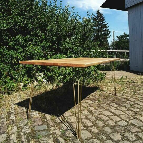 Moderner Esstisch aus Holz mit goldfarbenen Hairpin-Beinen, fotografiert auf einem gepflasterten Bereich vor einer grünen Hecke unter blauem Himmel.